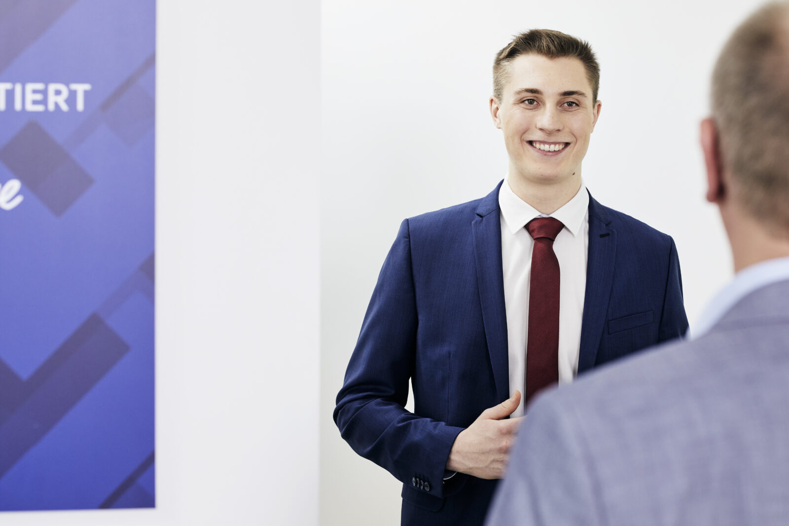 Junger Mann im blauen Anzug mit roter Krawatte lächelt im Gespräch.