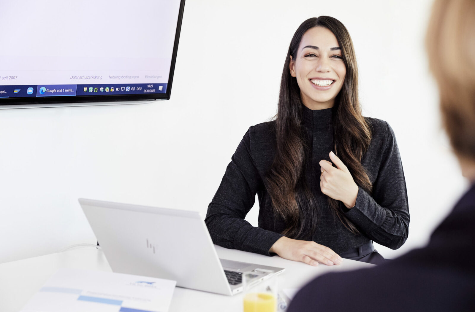 Frau lächelt im Gespräch an einem Tisch mit Laptop in einem Meetingraum.