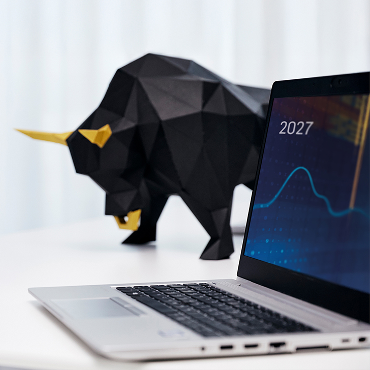 Ein geöffneter Laptop im Vordergrund zeigt ein Börsen-Chart mit der Jahreszahl 2027, während im unscharfen Hintergrund eine moderne, geometrische Skulptur eines schwarzen Bullen mit goldenen Hörnern steht – passend zur Branchenumfrage "AVD-Barometer 2027" von dwpbank und lemon.markets, die Finanzinstitute über die Einführung des Altersvorsorgedepots befragte.